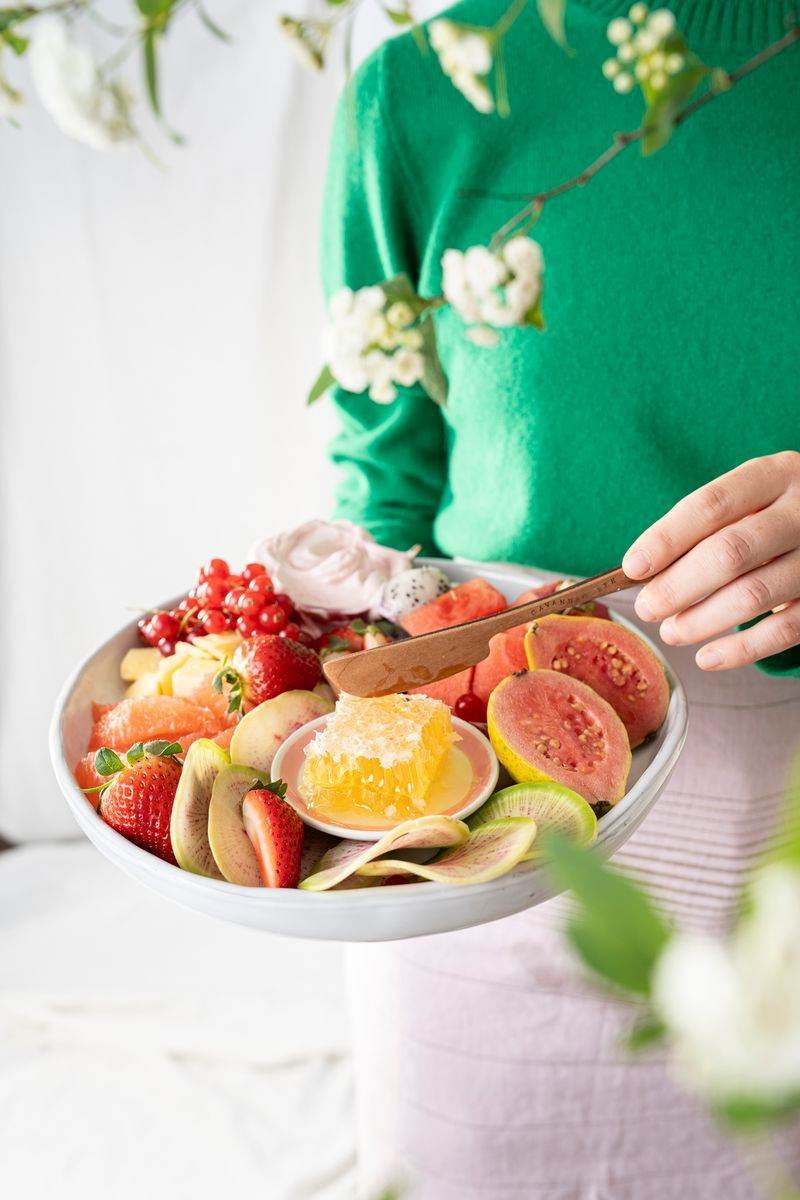 A woman surrounded by flowers holding a bowl of pink fruits and raw honeycomb.