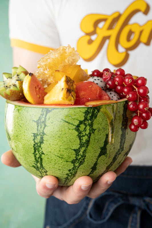 Person holding a watermelon that is cut in half and served with an assortment of fruit and raw honeycomb inside. 