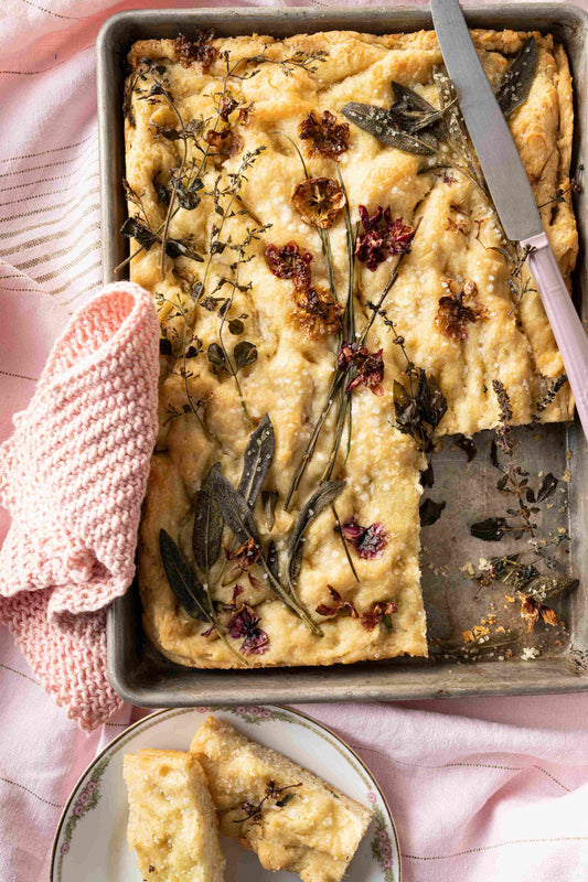 Flower focaccia on a baking tray with a pink knit blanket and butter knife. 