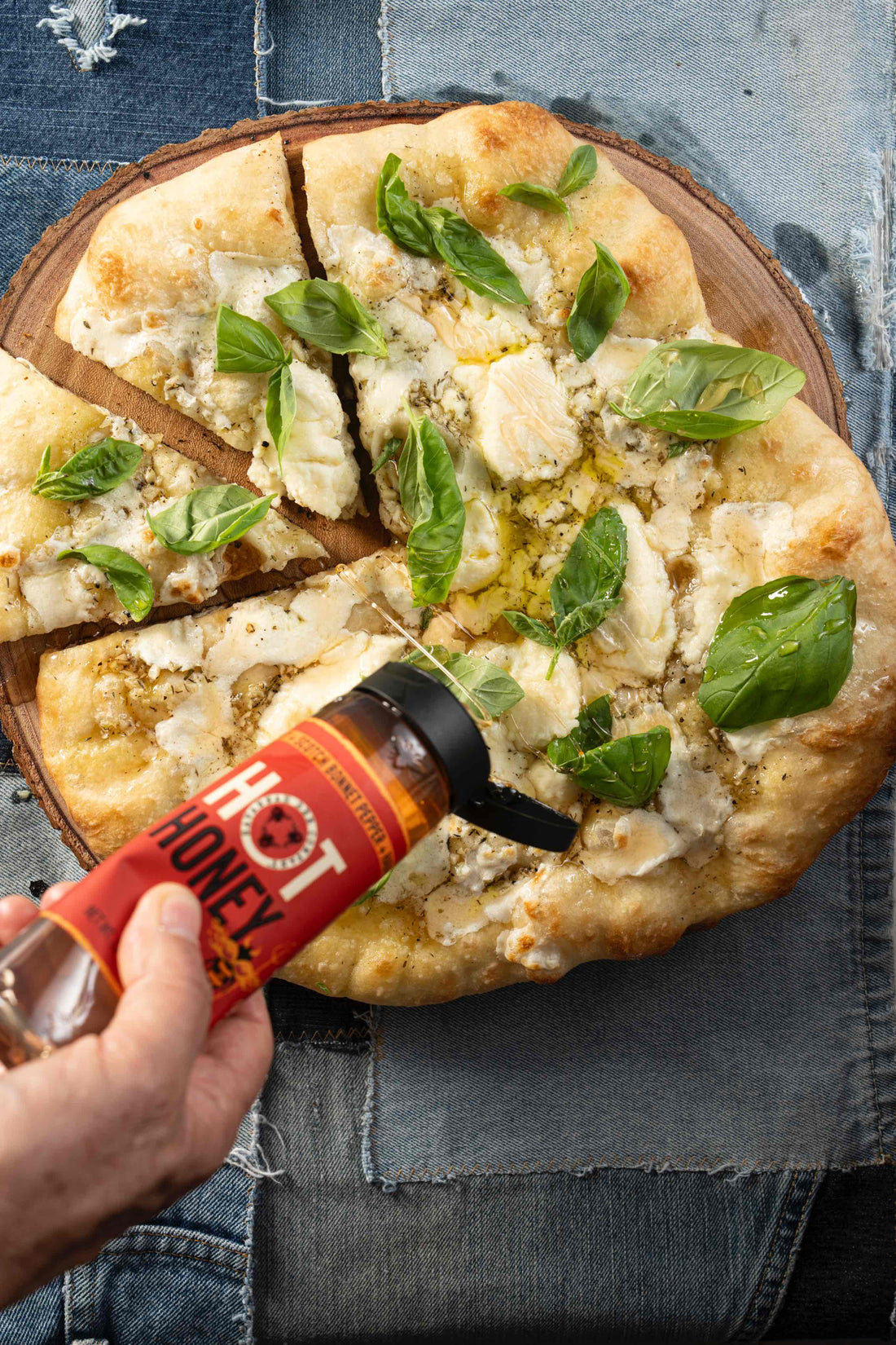 A rustic white pizza covered in olive oil and spices as well as cheese and basil sits on a wooden pizza board while being drizzled with Savannah Bee Company Hot Honey