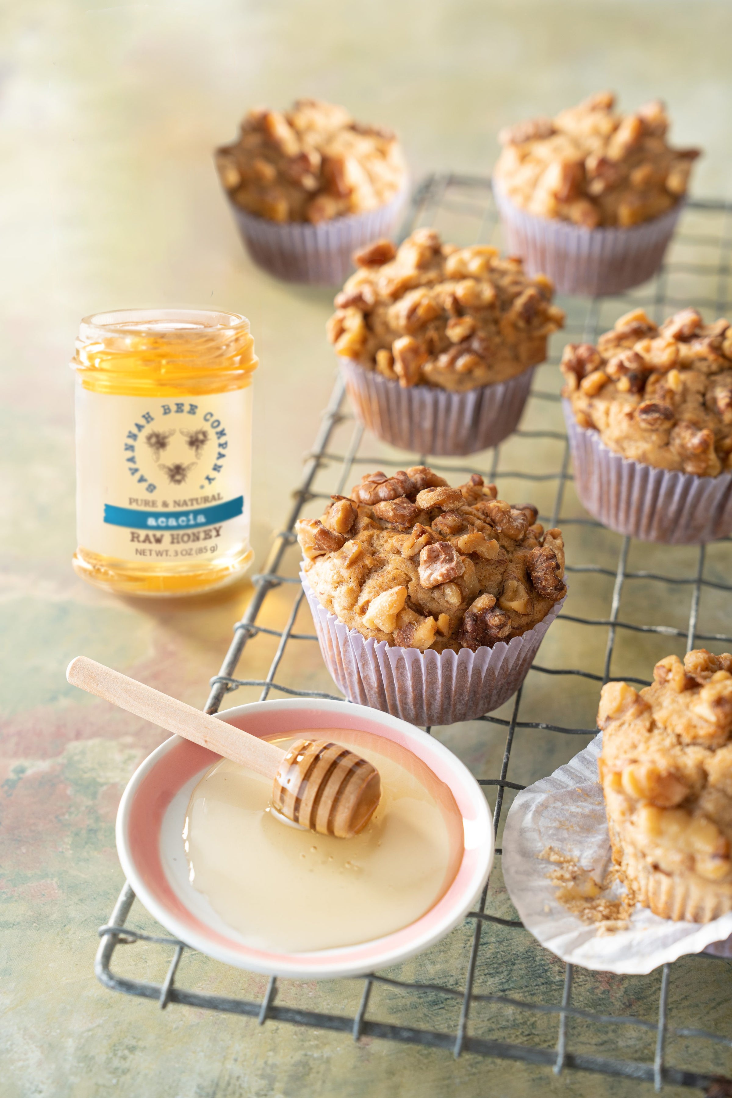 Mini honey dipper in a platter of honey next to a jar of acacia honey and a banana nut muffin.