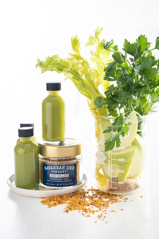 Bee pollen jar next to 3 celery wellness shots and a cup of ingredients for celery wellness shots.