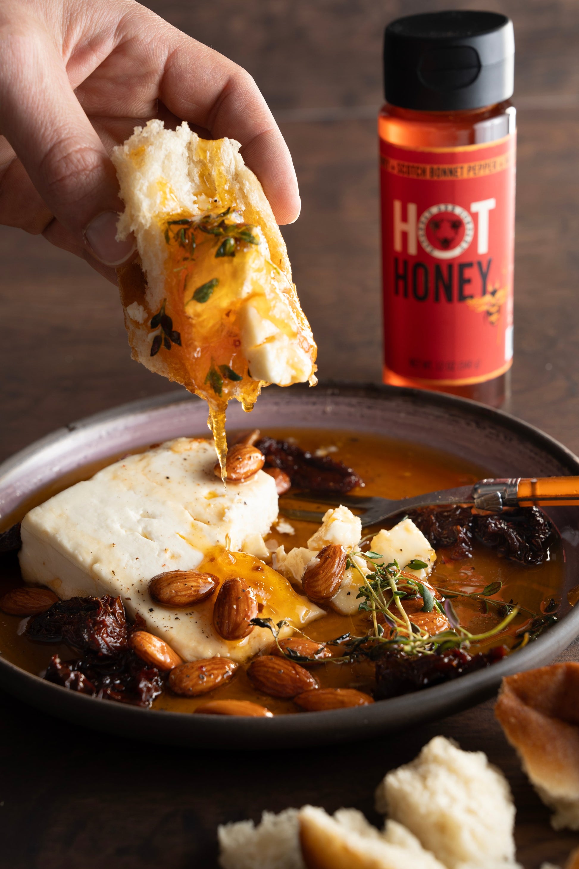 12 ounce hot honey squeeze bottle next to a plate of baked feta with almonds, hot honey, raisins and herbs.