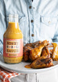 A model holding Savannah Bee Company mustard BBQ sauce bottle on a plate of chicken wings