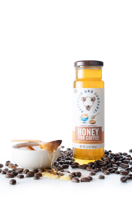 Honey for coffee in a glass jar next to a spoonful of honey and surrounded by coffee beans on a white background