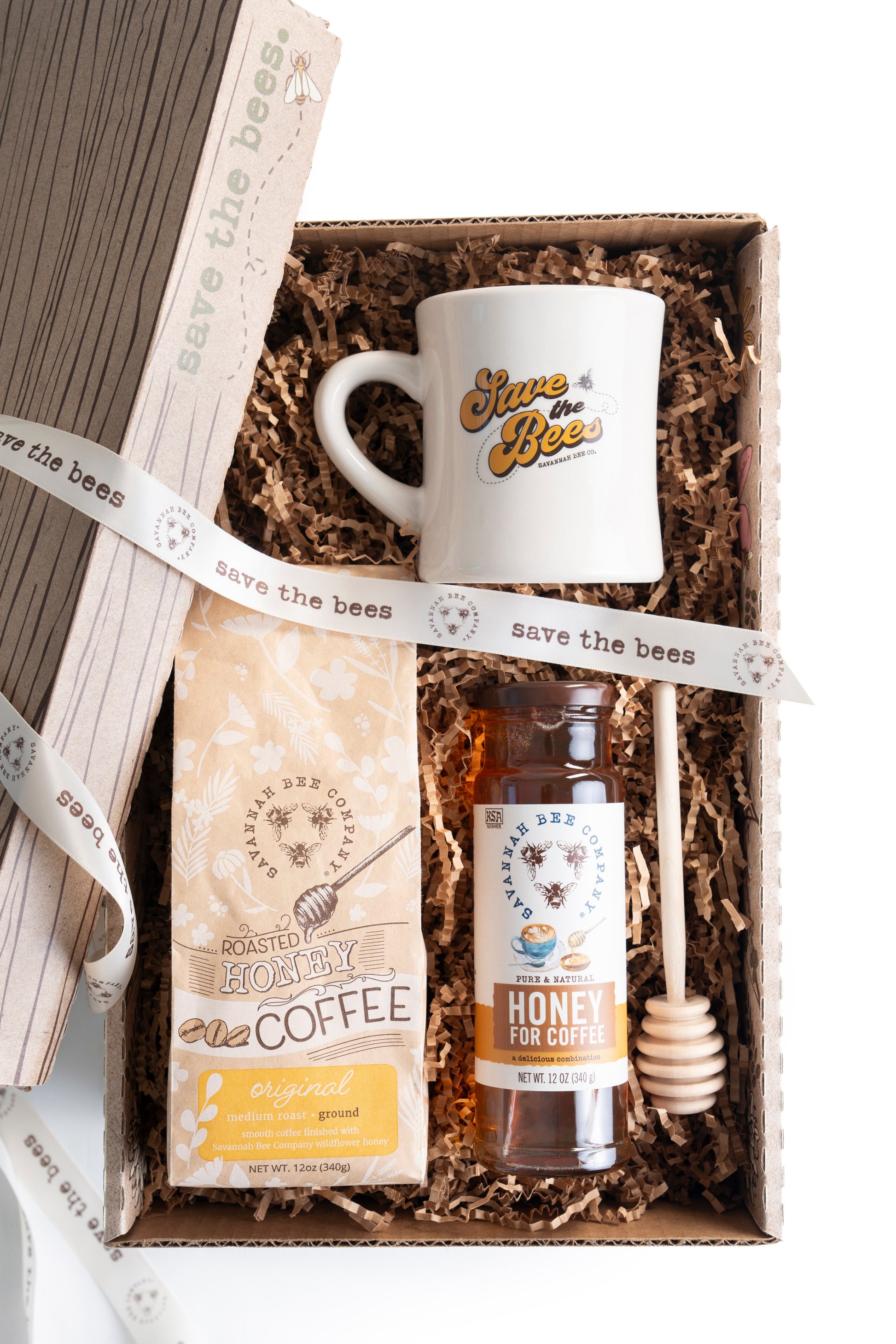 Overhead view of an open box with a save the bees diner mug ground honey roasted coffee bag honey for coffee and a honey dipper.