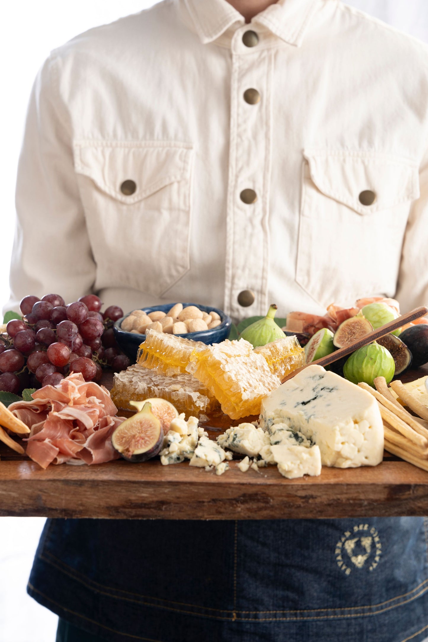 Cheese board held by someone, featuring grapes, nuts, blue cheese, figs, cured meats, and honeycomb.