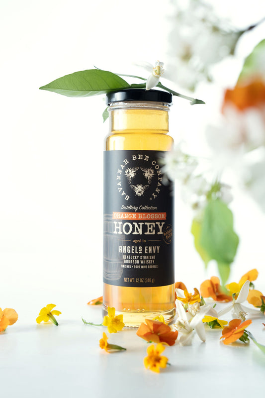 12 ounce Port Bourbon Barrel Aged Orange Blossom Honey  surrounded by orange blossoms against a white background.