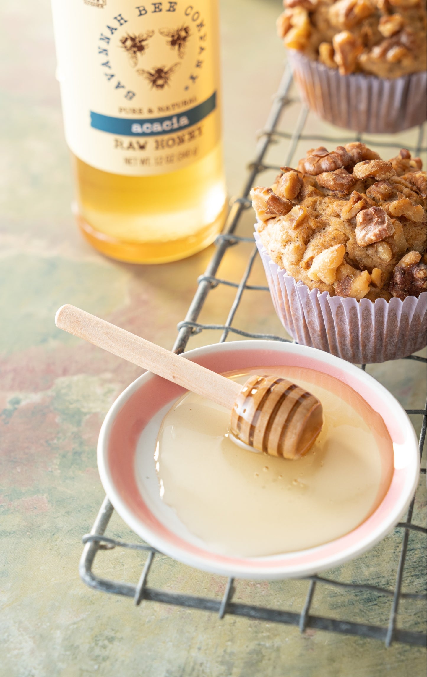 Mini honey dipper in a platter of honey next to a jar of acacia honey and a banana nut muffin.