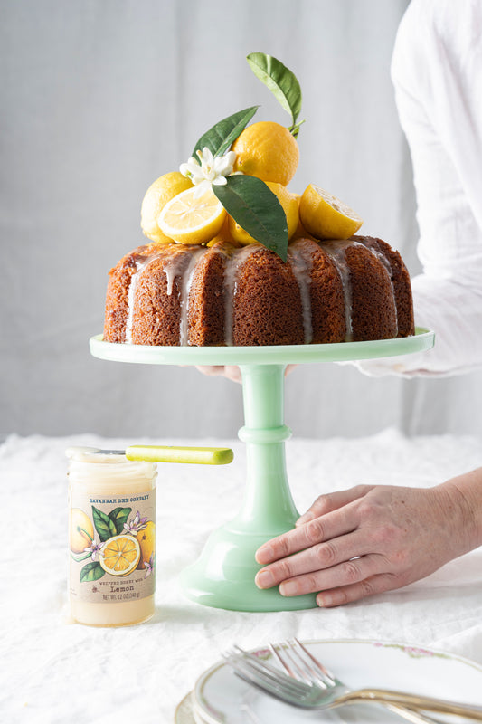 Lemon pound cake with lemons on top on a cake stand, next to whipped lemon honey.