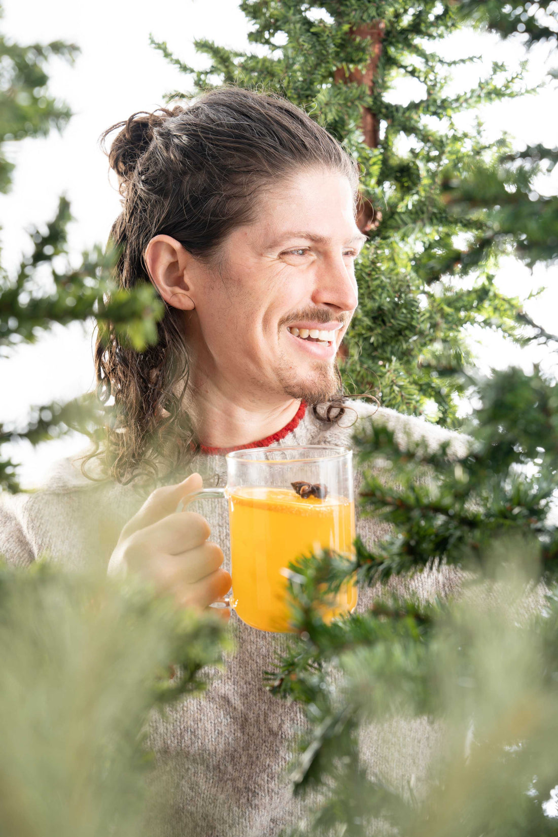 Man holding a drink surrounded by evergreen trees. 