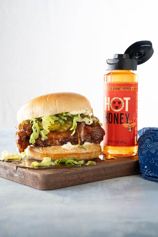 A hot honey chicken sandwich with lettuce and pickles on a wooden cutting board next to a 12 ounce squeeze bottle of Savannah Bee Company Hot Honey.