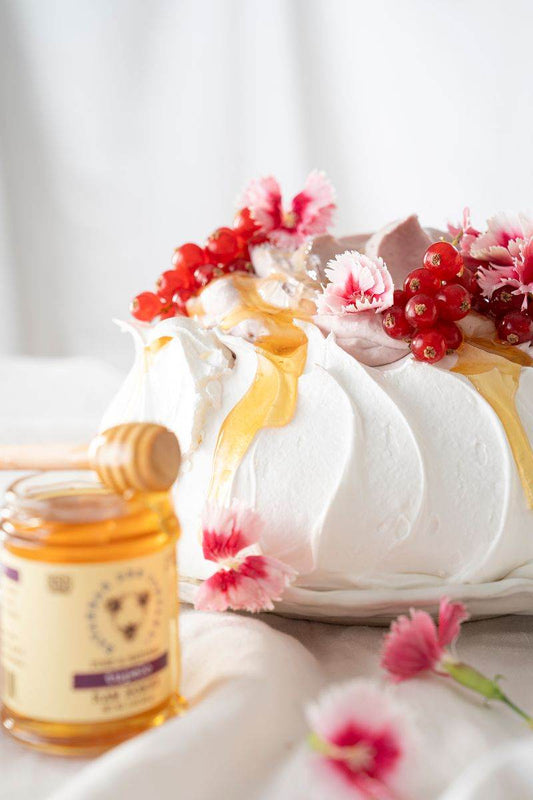 Blackberry pavlova covered with edible flowers, red berries, and honey, next to a 3 ounce jar of tupelo honey and a honey dipper.