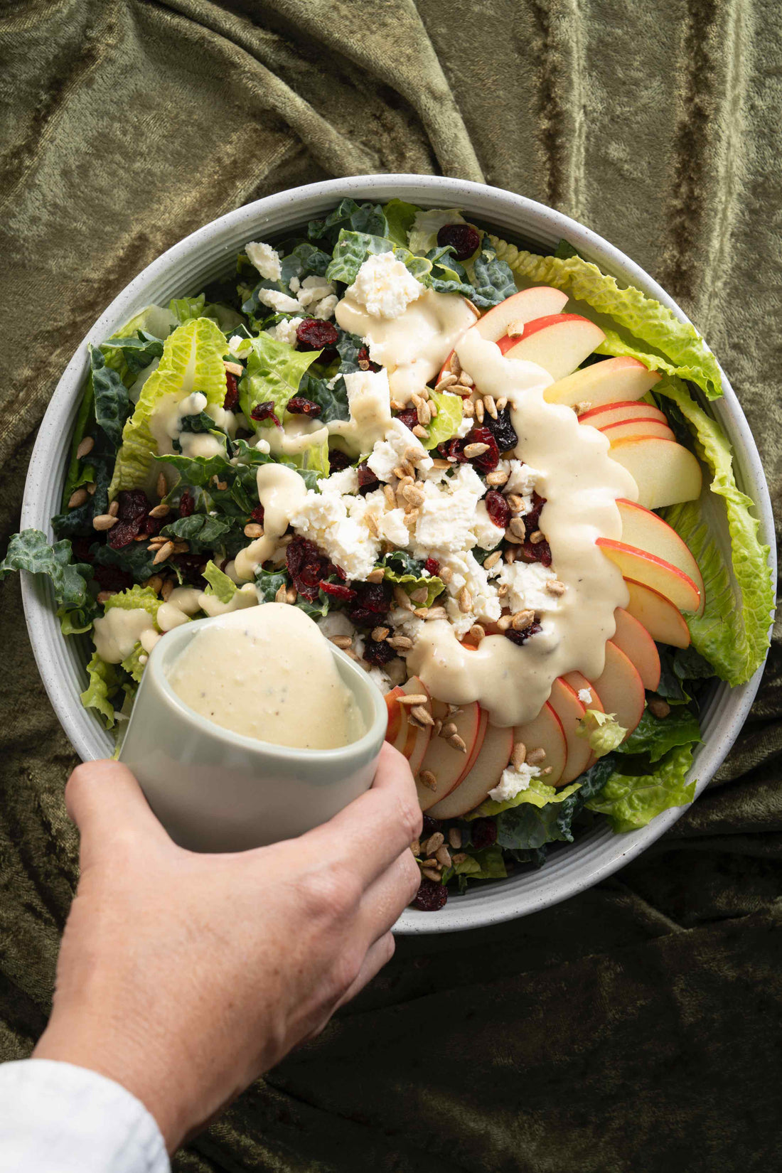 A Creamy Honey Dressing is drizzled over a large bowl of salad garnished with Apples, Feta Cheese, Dried Cranberries, and salted Sunflower Seeds.