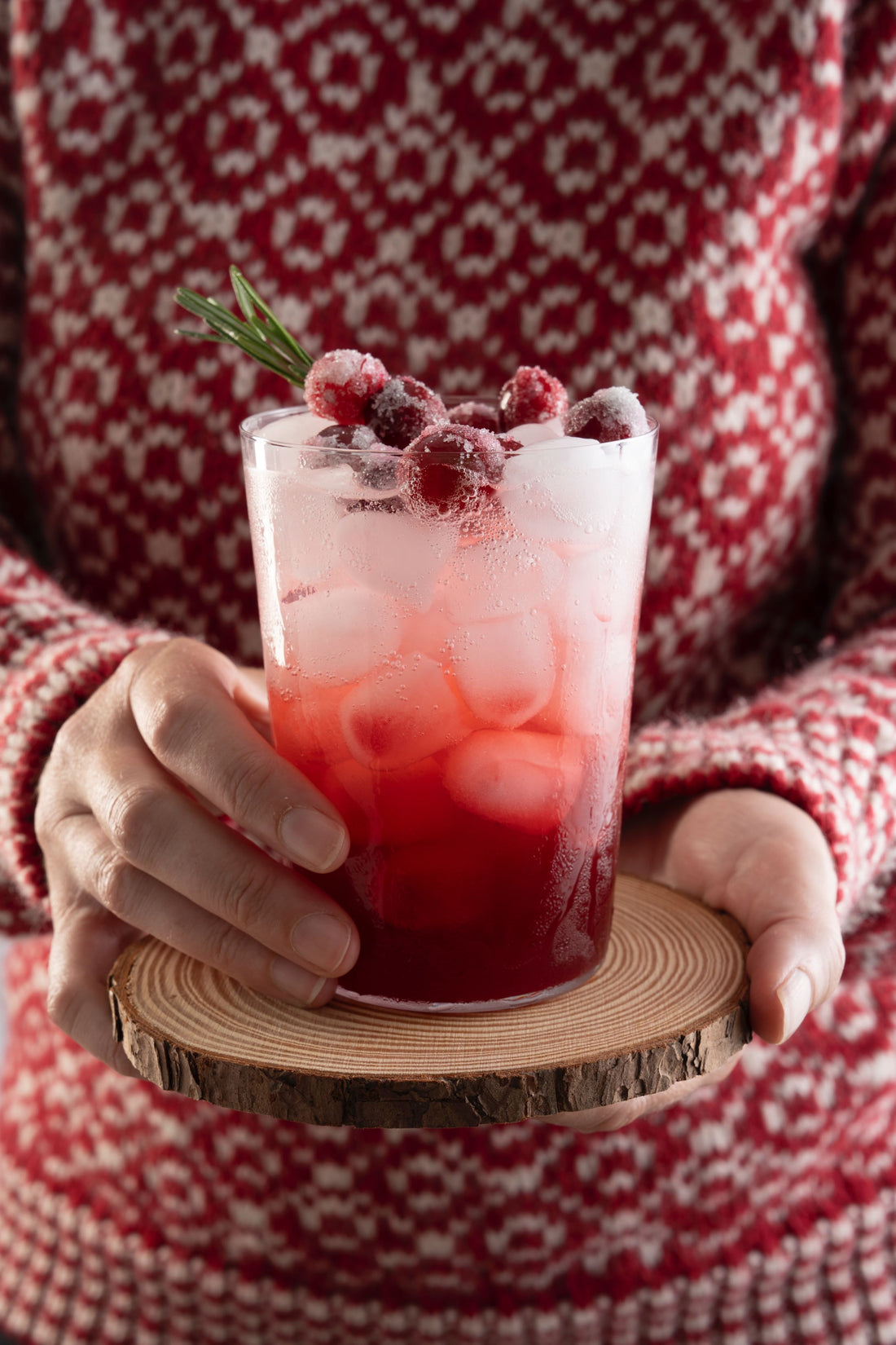 A woman in a red and white patterened wool sweater holds a small wooden coaster with a clear glass containing a red and white iced effervescent shrub cocktail garnished with rosemary and sugared cranberries.