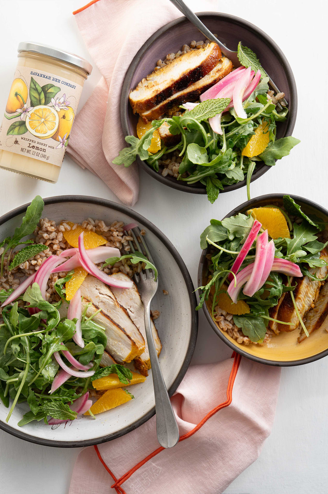 Three bowls of Honey Chicken Grain Salads sit on a white table with a jar of Savannah Bee Company Whipped Honey with Lemon. In the bowl you can see sliced chicken breast, arugula, mandarin oranges, farro and pickled red onion.