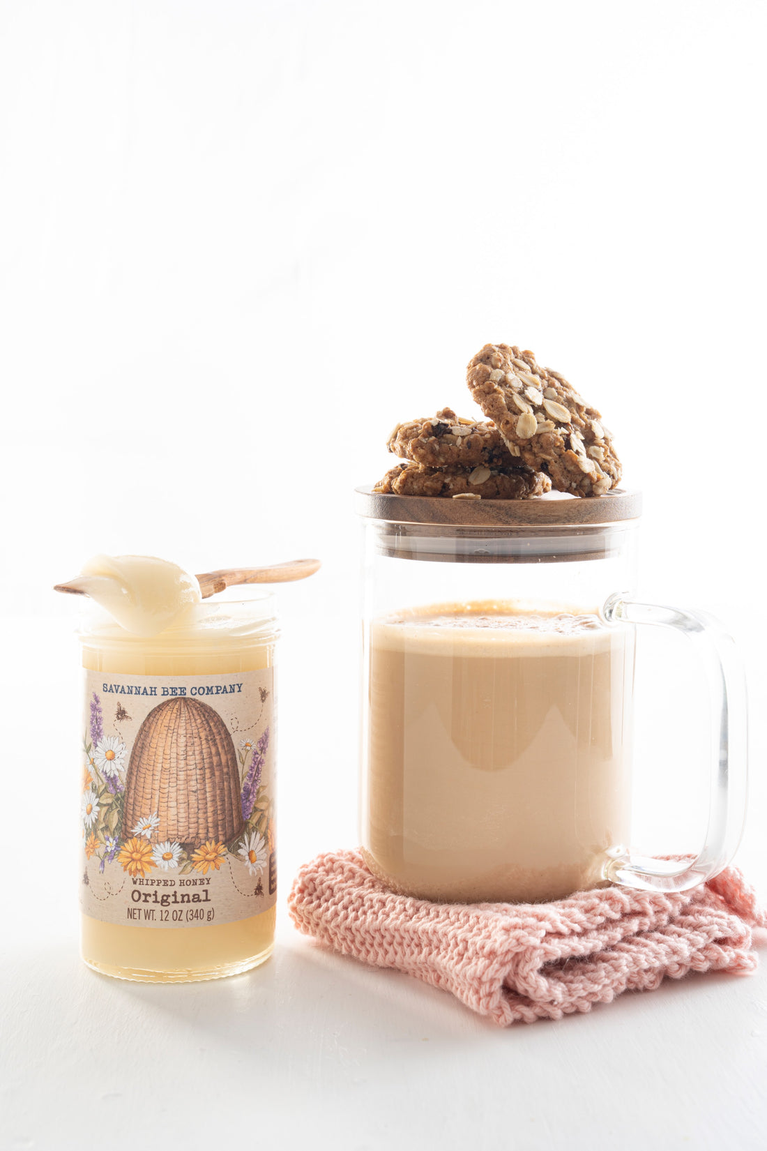 A single glass mug filled with a creamy latte capped with a wooden lid and three small oatmeal cookies, sitting on a folded pink towel next to a jar of Original Whipped Honey. 