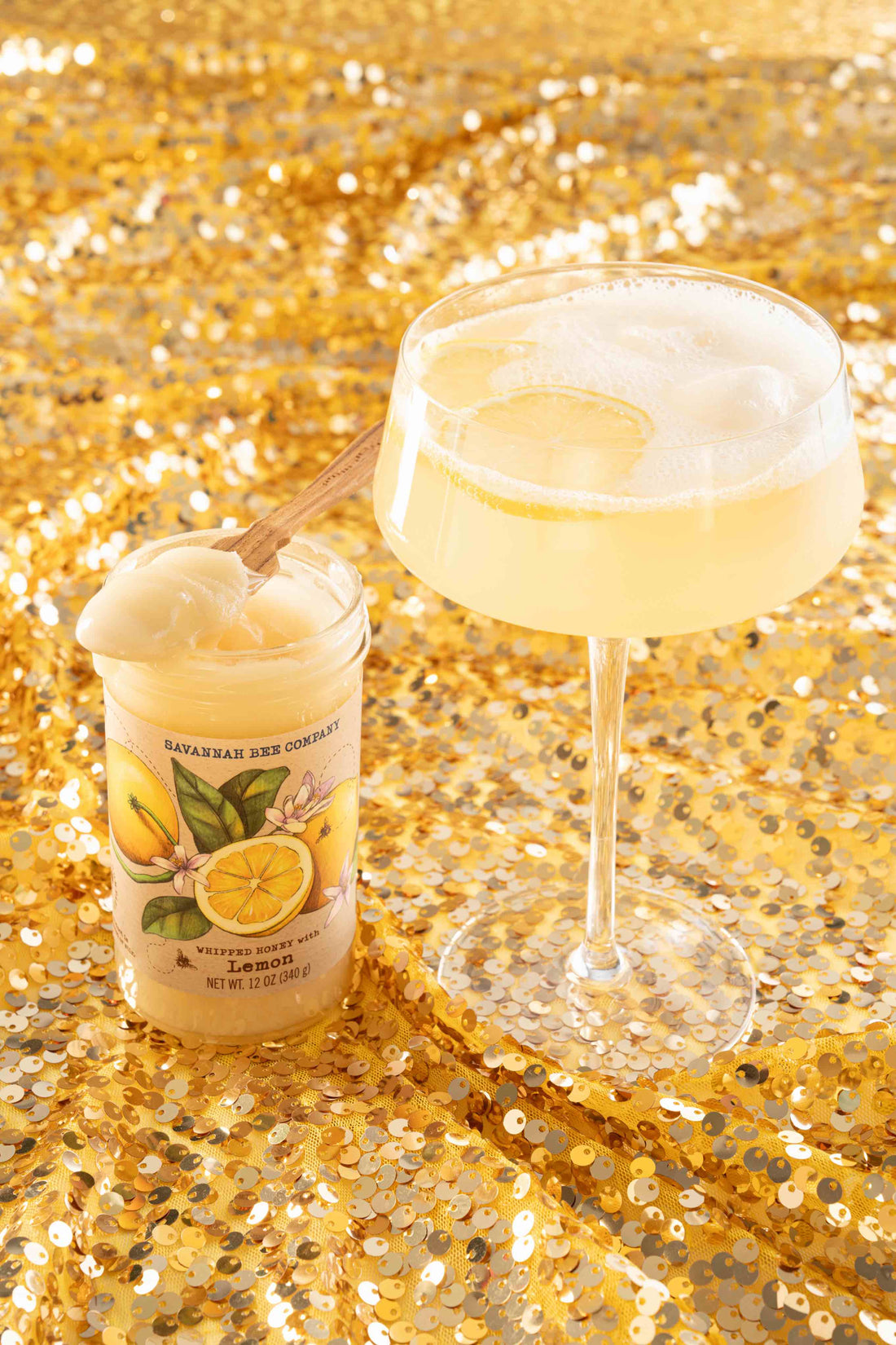 A champagne coupe is full of a fizzing lemon cocktail with a dollop of ice cream and standing next to a jar of whipped honey with lemon