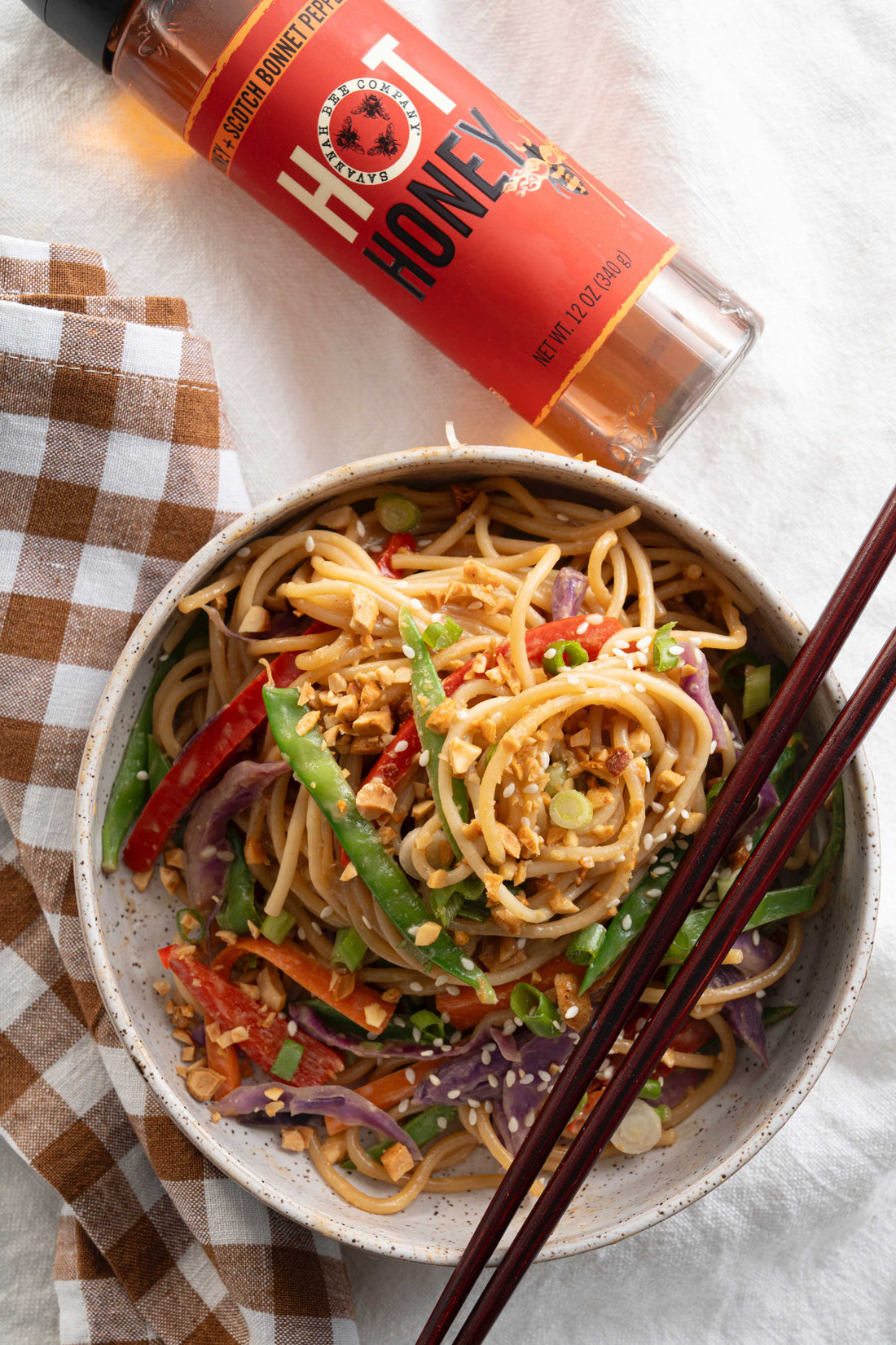 An overhead photo of a bowl of Spicy Peanut Noodles with fresh vegetables served with a squeeze bottle of Savannah Bee Company Hot Honey and a pair of wooden chopsticks on a white tablecloth.