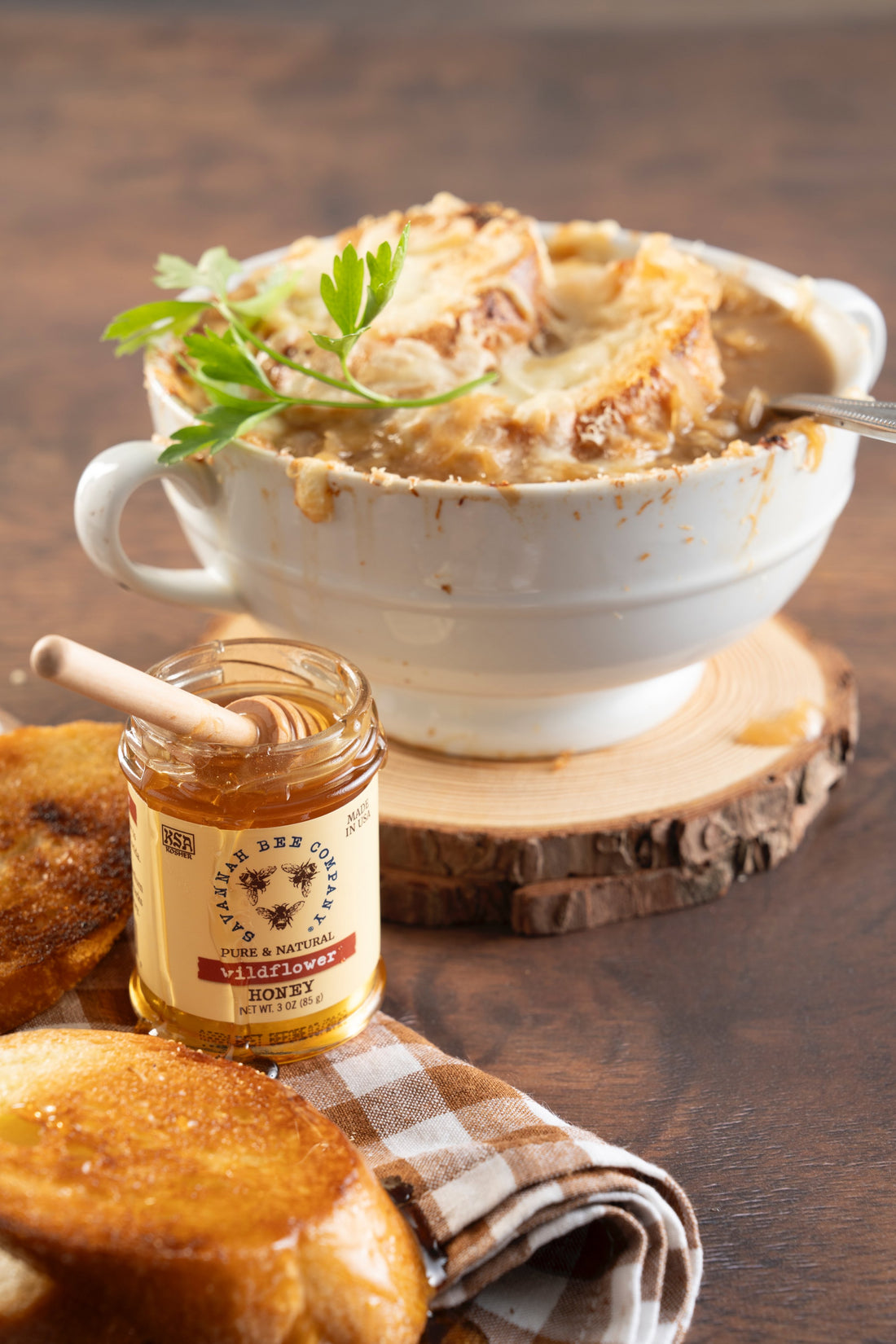 A heaping white bowl of french onion soup topped with melted cheese and thick slices of tasted baguette on a wooden trivet next to a small jar of savannah bee company wildflower honey.