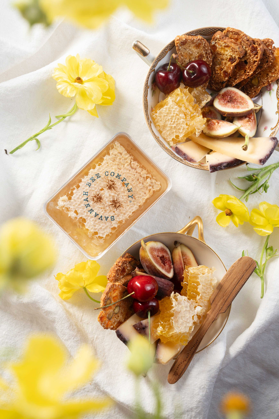 Two teacups filled with dried fruit crackers, chunks of raw honeycomb, halved figs, pitted cherries and slices of merlot-washed cheese are set on a white tablecloth with yellow flowers scattered around a container of Savannah Bee Company Raw Honeycomb.