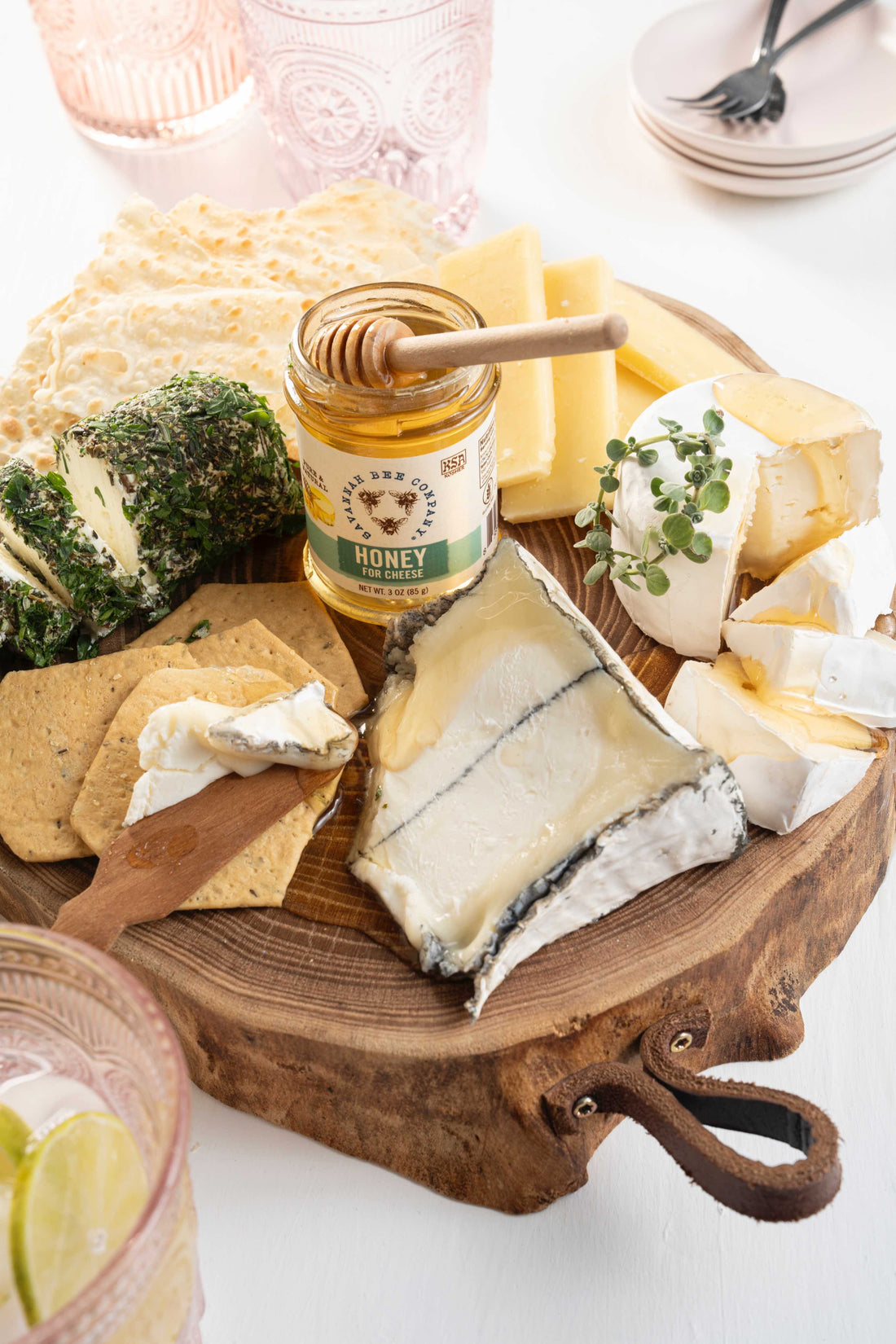 On a wooden board several cheeses circle a small jar of Savannah Bee Company Honey for Cheese. The cheeses are humboldt fog, herb crusted goat cheese, aged cheddar, and brie wedges. Crackers are being spread with wooden spreading knives. 