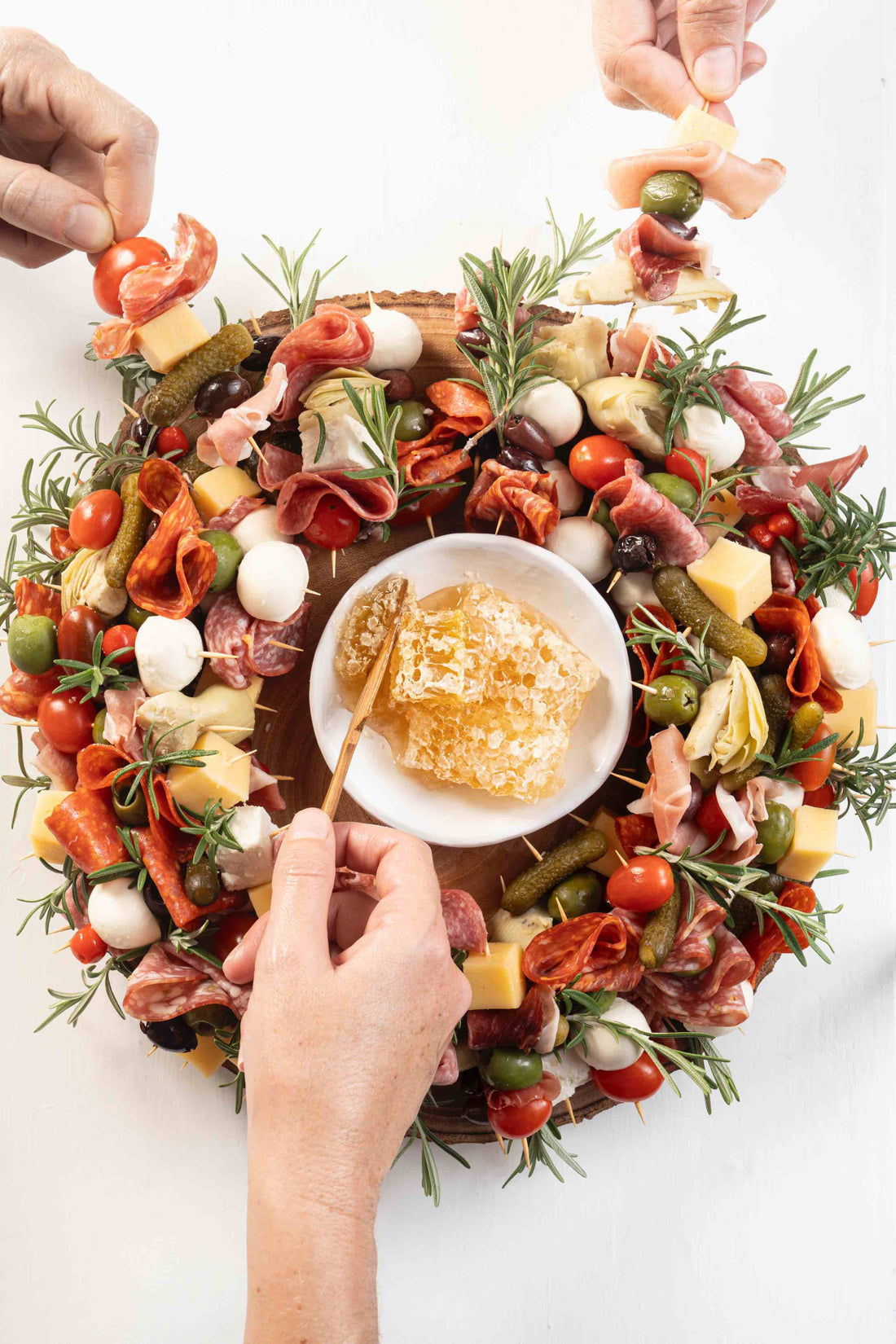 Three friends reach to grab a prepared charcuterie skewer from a platter fashioned to look like a holiday wreath; in the center of the wreath is a bowl of raw honeycomb with a teak honeycomb knife cutting through the honeycomb.