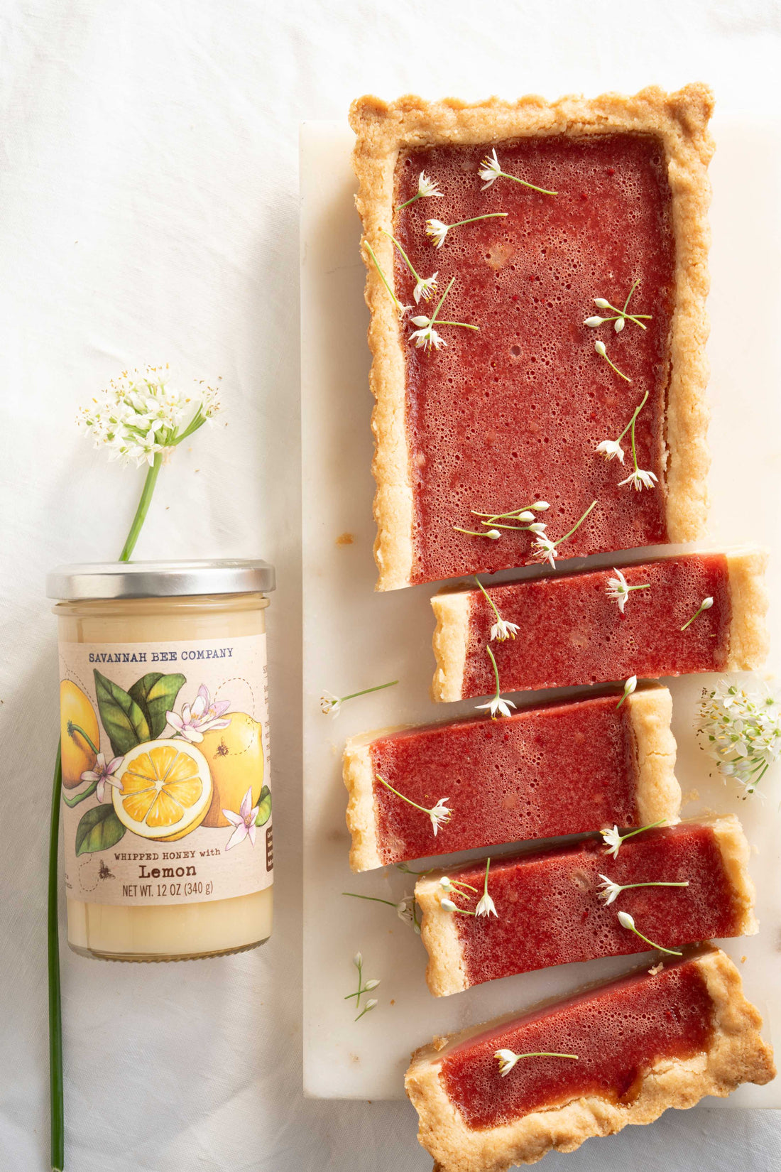 A long thin rectangular tart with a shortbread crust and a deep red cranberry filling is sliced into one inch pieces and decorated with small white edible flowers on a white tray next to a jar of Savannah Bee Company Whipped Honey with Lemon. 