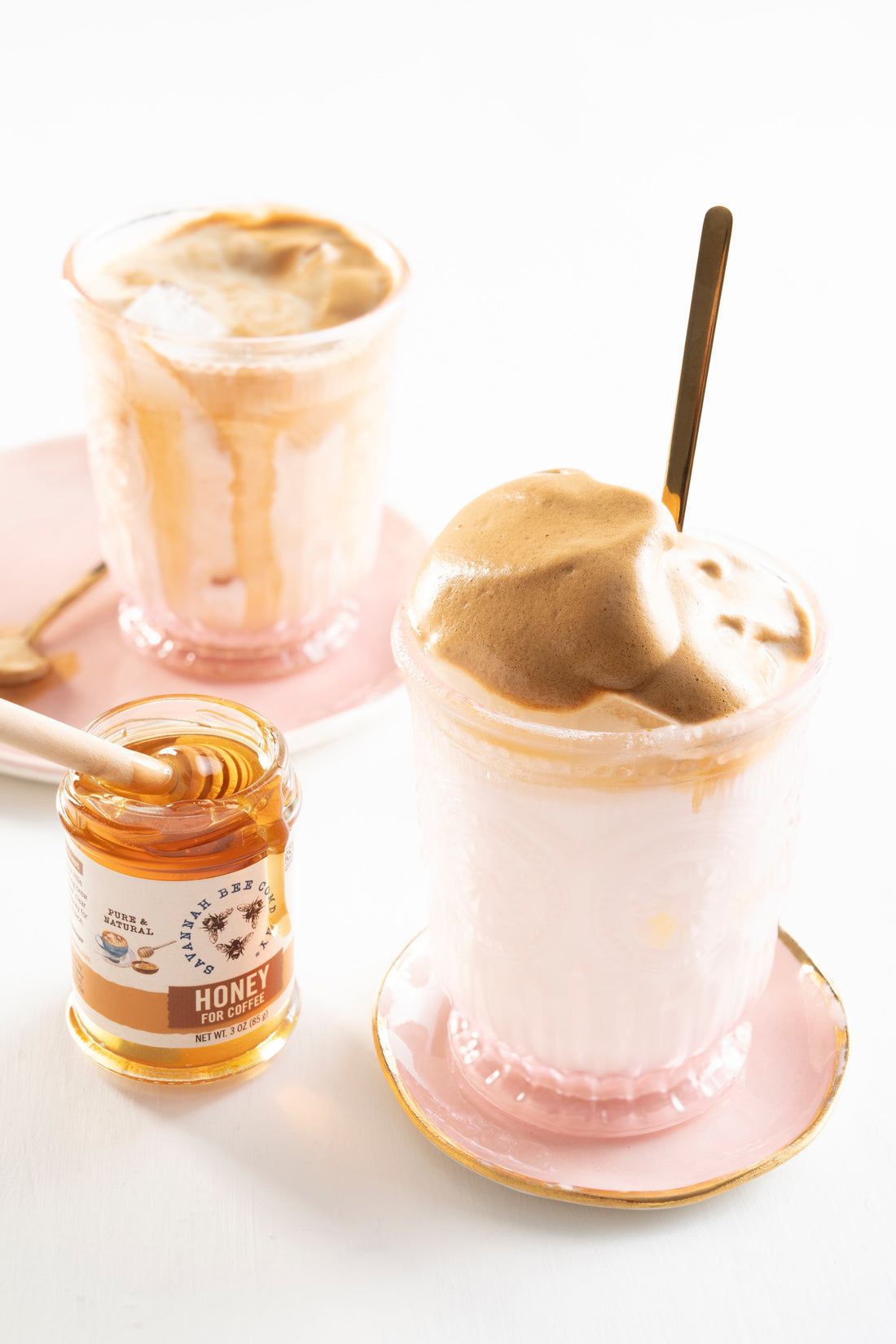 Two glasses on pink saucers are filled with milk topped with voluminous and cloud like whipped honey coffee next to a jar of Savannah Bee Company Honey For Coffee.