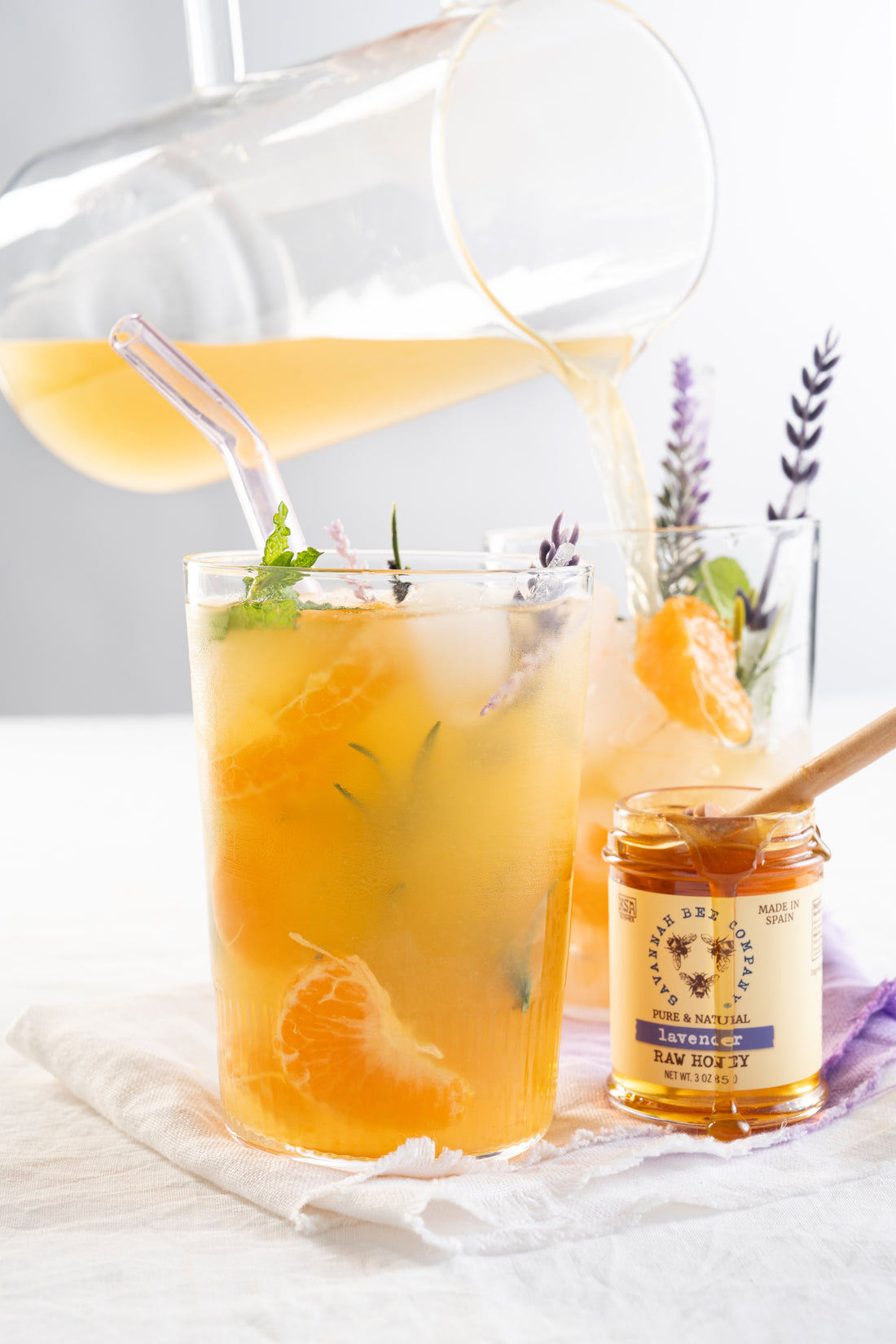 A pitcher of Lavender Lemonade coctail is being poured into two glasses with lavender ice cubes on a white table with white cocktail napkins next to a small jar of Savannah Bee Company Lavender honey.