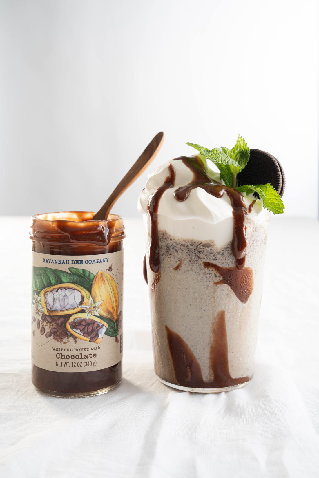 A clear frosted glass on a white table cloth is filled with a cookies and cream shake, topped with whipped cream, an oreo cookie, a mint sprig and drizzled with chocolate whipped honey from a jar of chocolate honey sitting next to the glass.