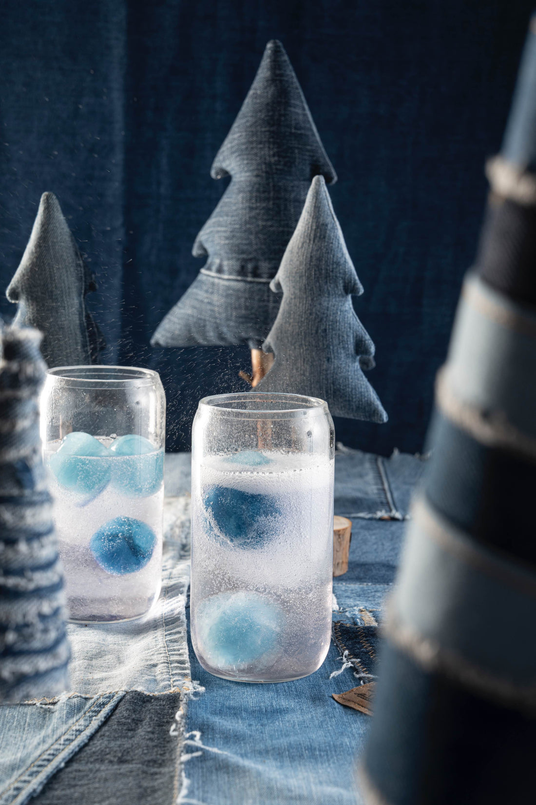 Two classes of an effervescent clear drink with large spherical blue ice cubes sit on a table covered in a denim table cloth with Christmas trees made of denim. 