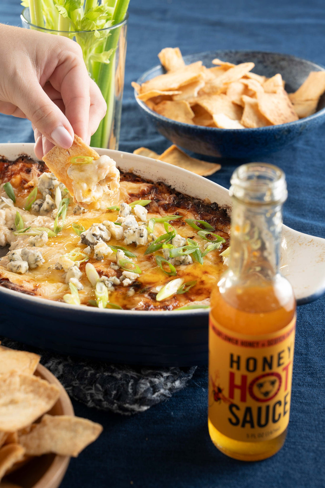 A hand dips a pita chip into a casserole dish of Buffalo chicken dip next to a bottle of Savannah Bee Company Honey Hot Sauce.