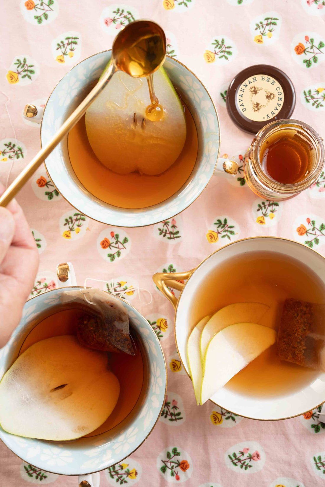 Three vintage teacups of tea with slices of pear as garnish sit on a table with a spoonfull of honey dropping into one of the tea cups.