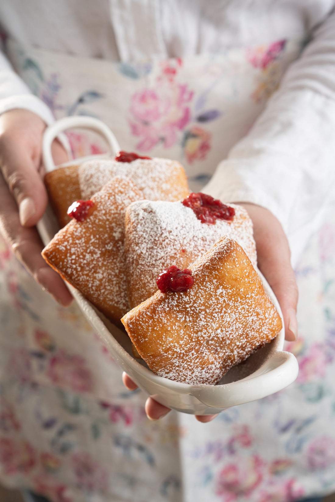 A woman in a flora apron holds a small tray with four jam filled square donuts lightly dusted with powdered sugar. 