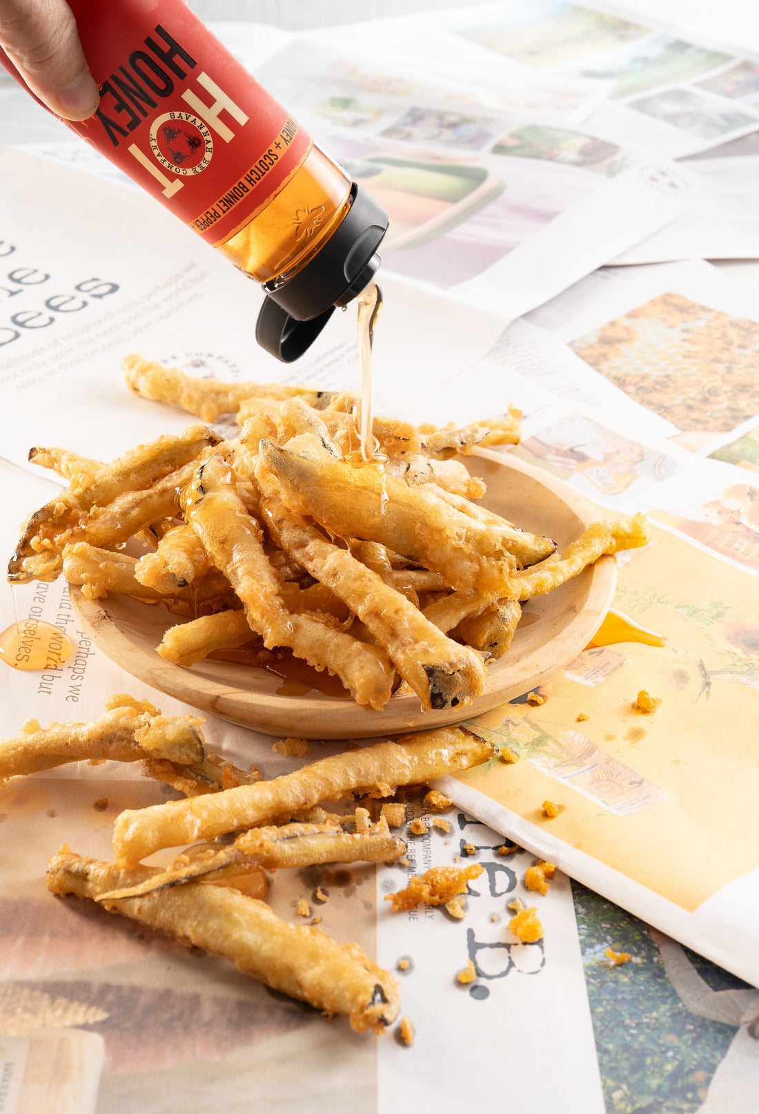 Someone drizzles Savannah Bee Hot Honey over a bowl of Eggplant Fries on a table covered with copies of the savannah bee company newspaper.