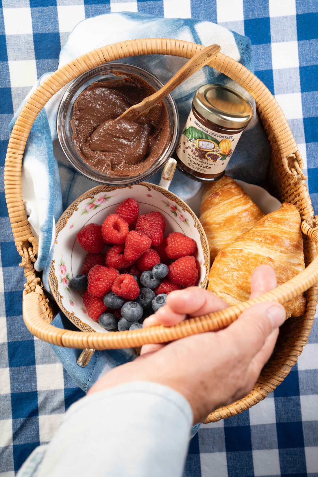 A picnic basket on a blue gingham blanket is filled with hazelnut spread, croissants, a teacup full of raspberries and blueberries and a small jar of Savannah Bee Company Whipped Honey with Chocolate.