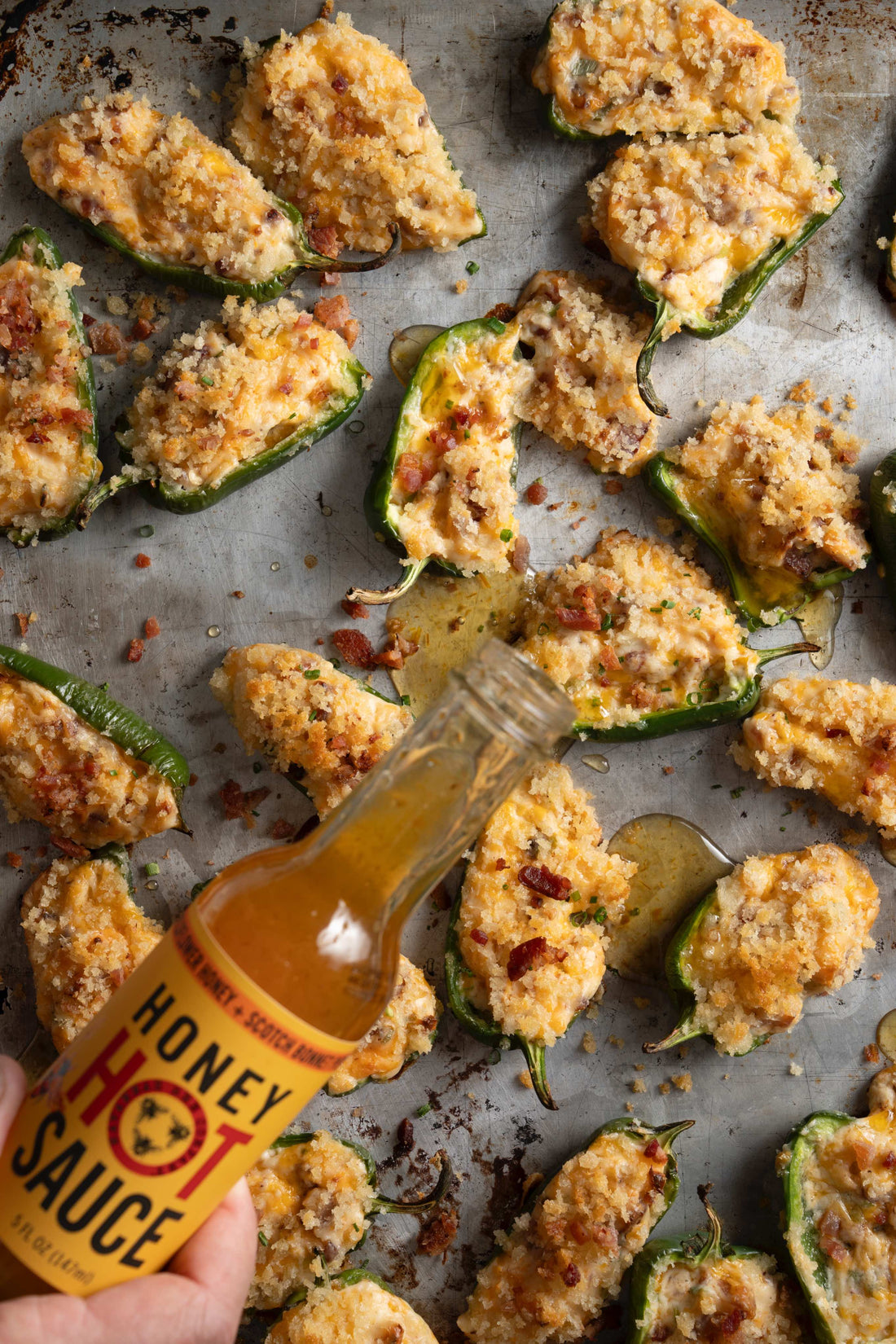 A bottle of Savannah Bee Company Honey Hot sauce pouring onto a baking sheet full of halved jalapeños stuffed with a cheesy filling.