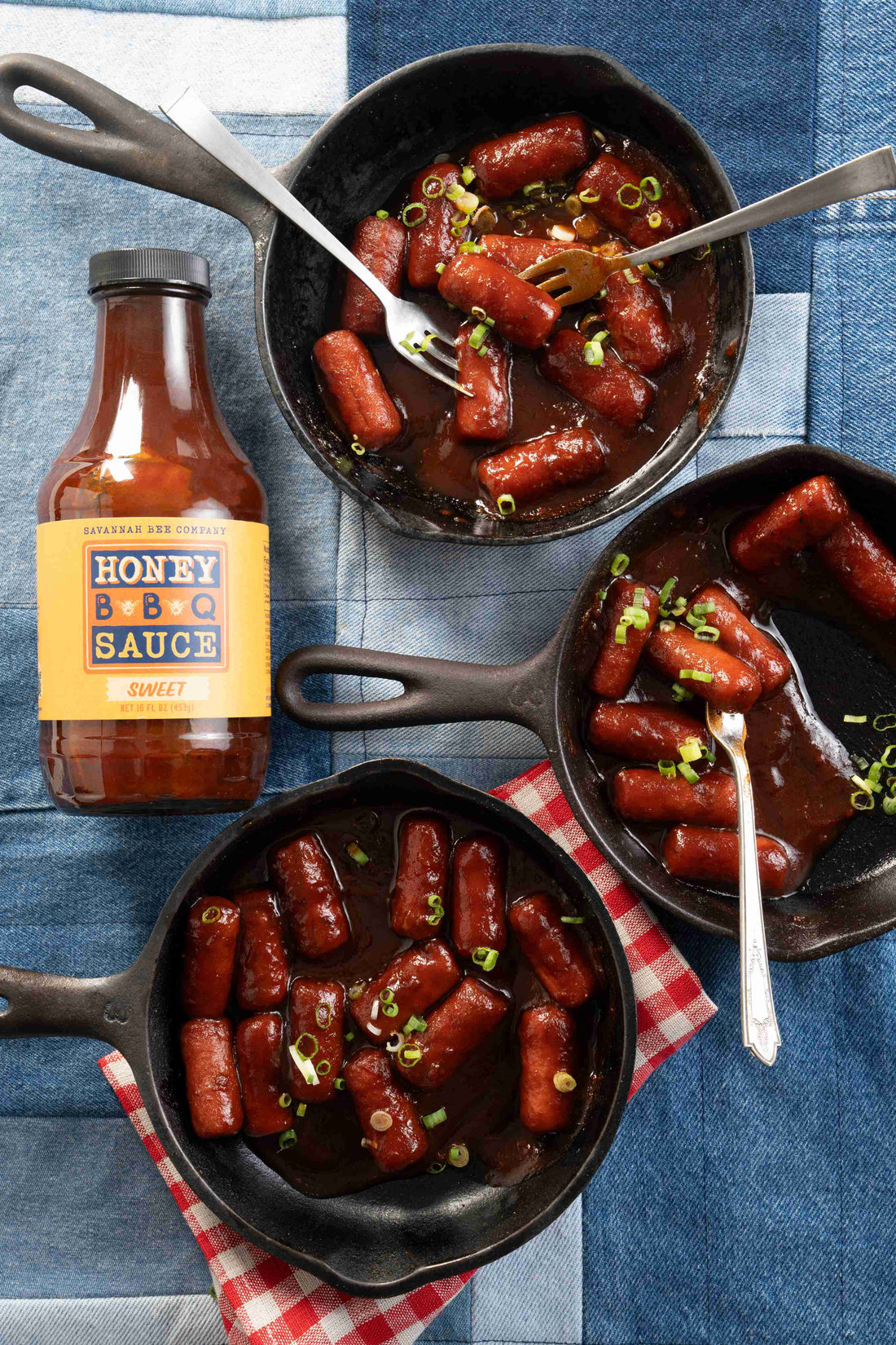 Three small cast iron pans filled with little smokies on a denim patchwork table cloth with a red gingham napkin next to a bottle of savannah bee company Honey BBQ Sauce.