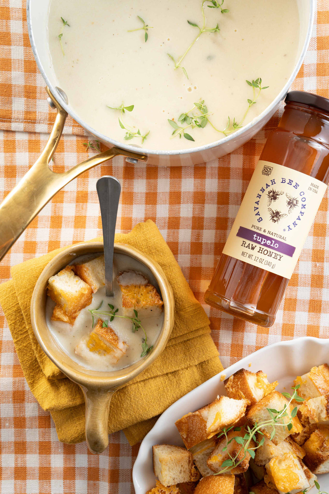 A bowl of soup with croutons and a garnish of thyme on a checkered table cloth next to a jar of Savannah Bee Company Tupelo honey and a large white pot of parsnip soup.