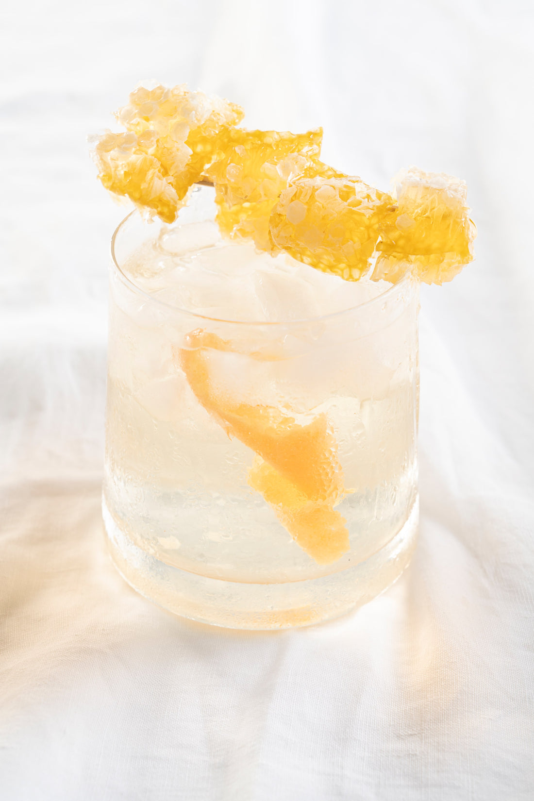 A single clear glass with a clear iced beverage is garnished with large chunks of honeycomb and  a pink grapefruit twist.