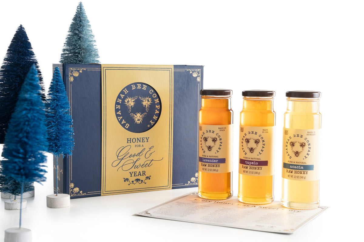 Three twelve ounce jars of honey next to the navy blue gifting box and multiple navy blue bottle brush trees.