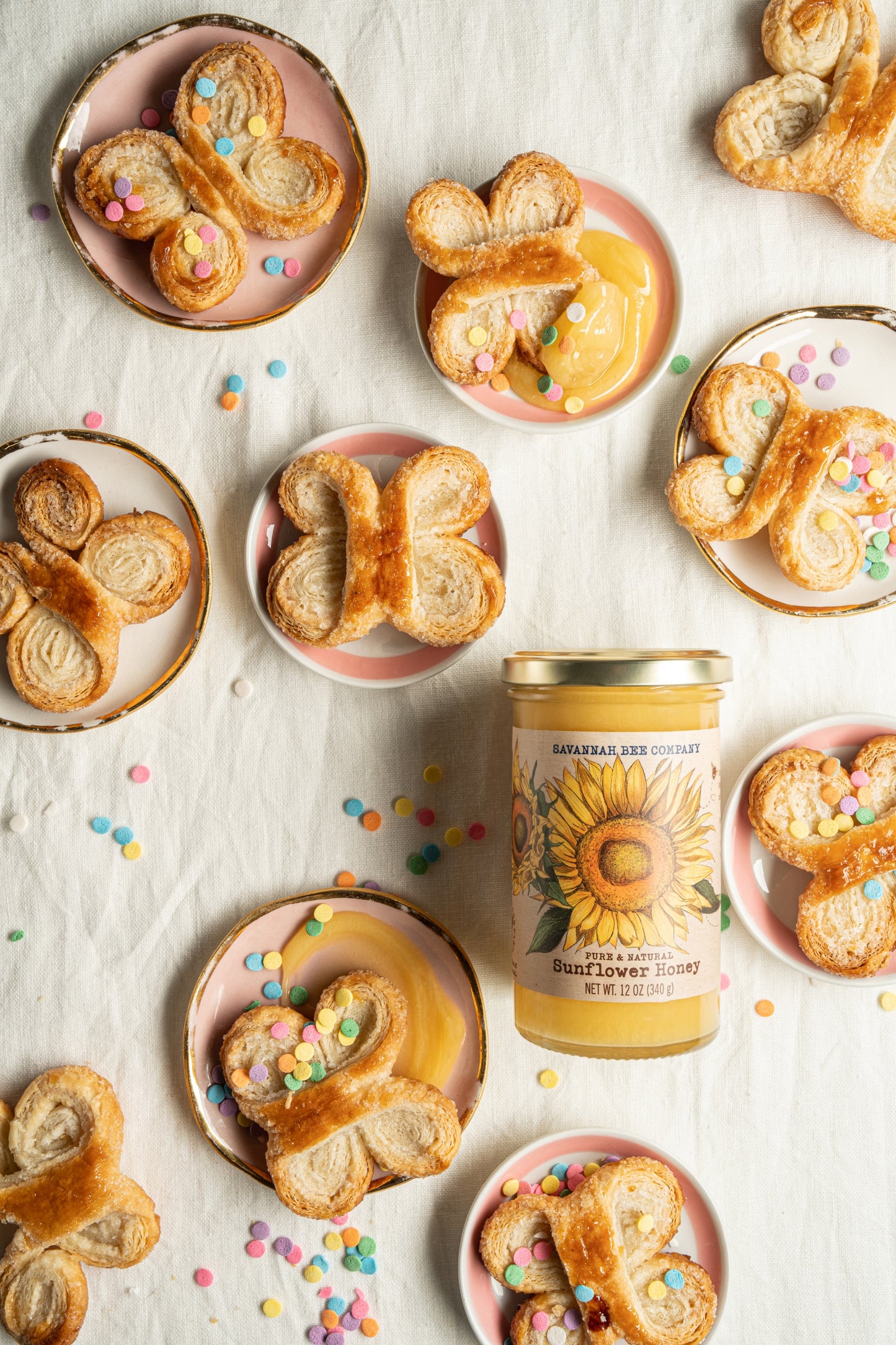 Butterfly shaped cookies on small plates with multicolored sprinkles next to a 12 ounce jar of sunflower honey.