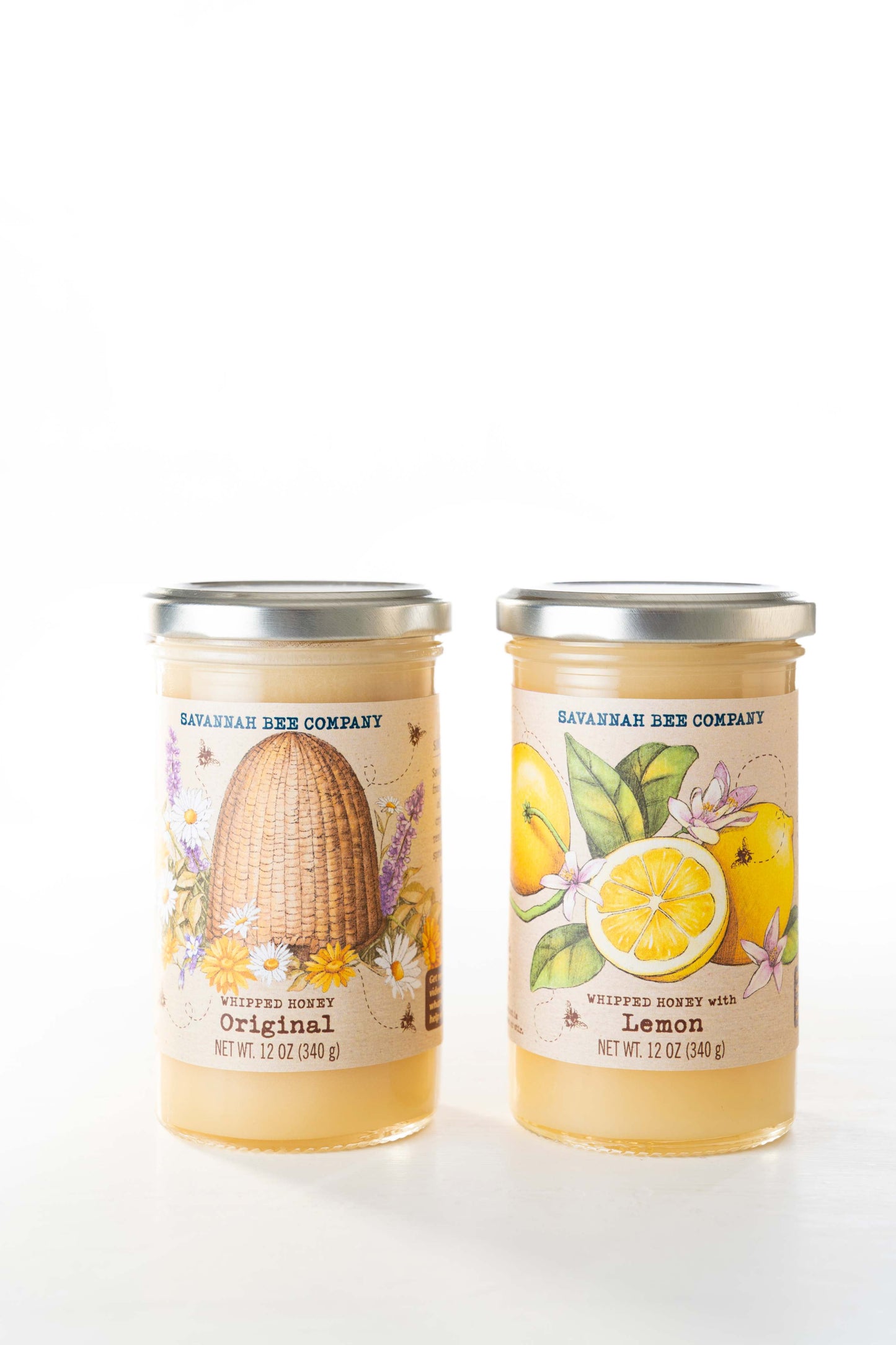 Two jars of whipped honey sit in a white space; the jar on the left is original whipped honey with a straw bee hive on the label, the jar on the right is lemon whipped honey with lemons and and lemon leaves on the label.
