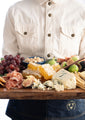 Cheese board held by someone, featuring grapes, nuts, blue cheese, figs, cured meats, and honeycomb.