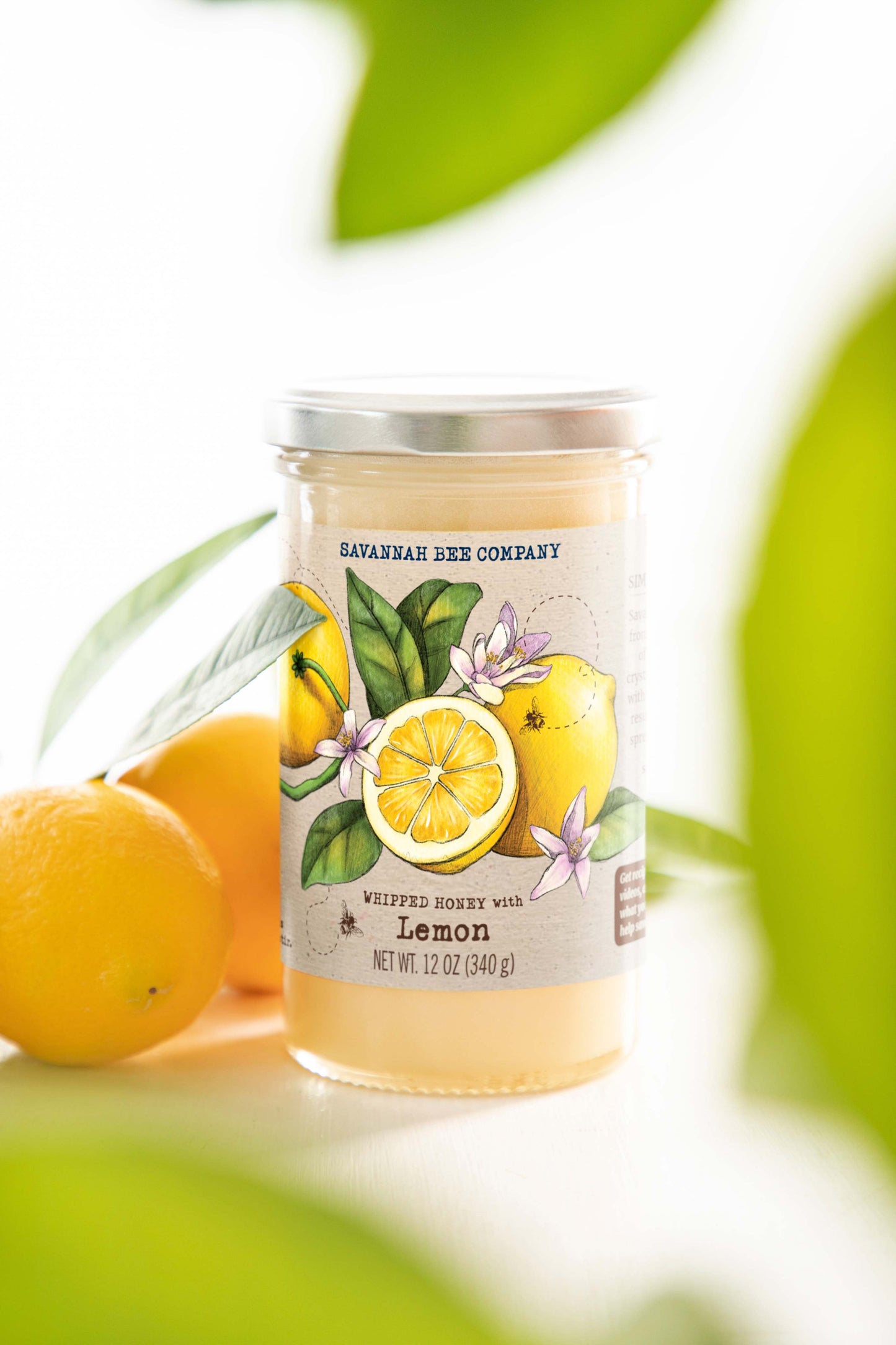 A jar of whipped honey with lemon sits on a white table with fresh lemons and lemon leaves. 