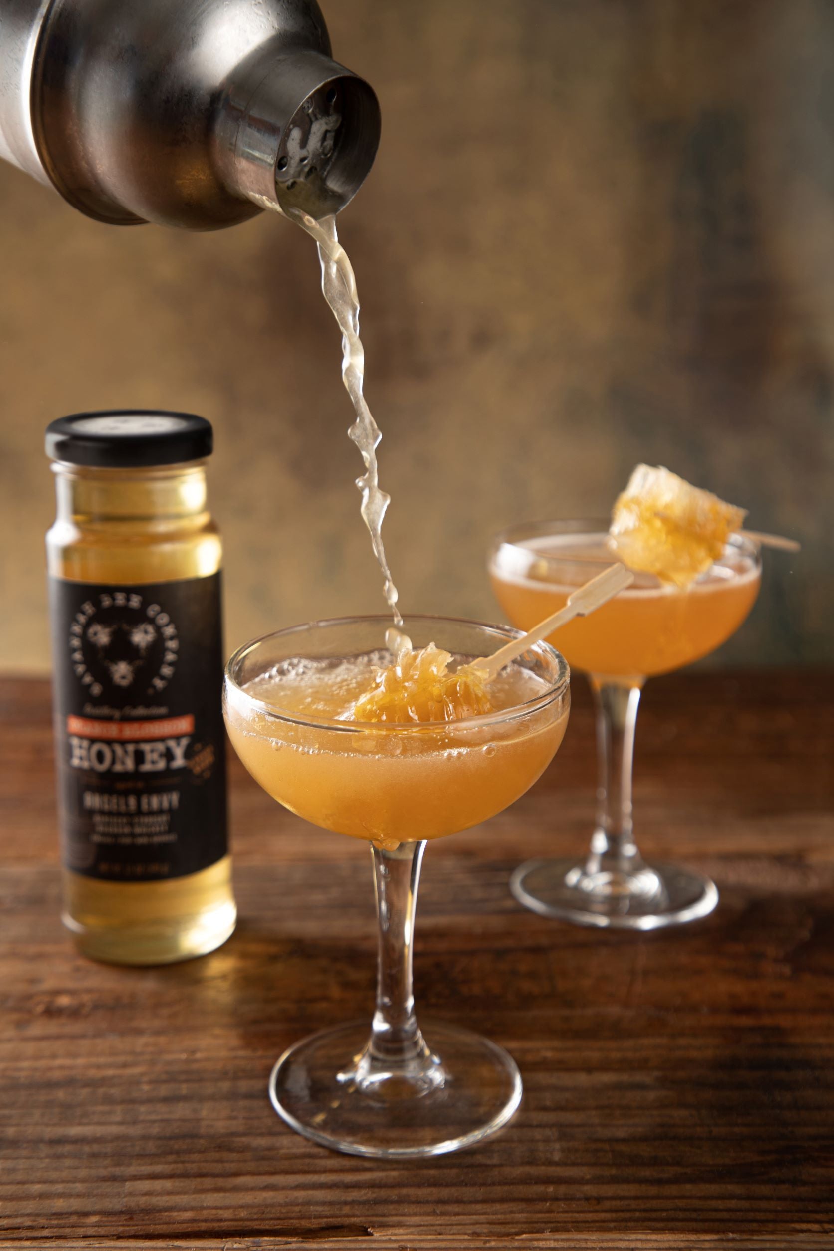 Two bourbon cocktails being poured from a shaker into cocktail glasses with a honeycomb garnish next to a 12 ounce Port Bourbon Barrel Aged Orange Blossom Honey jar.